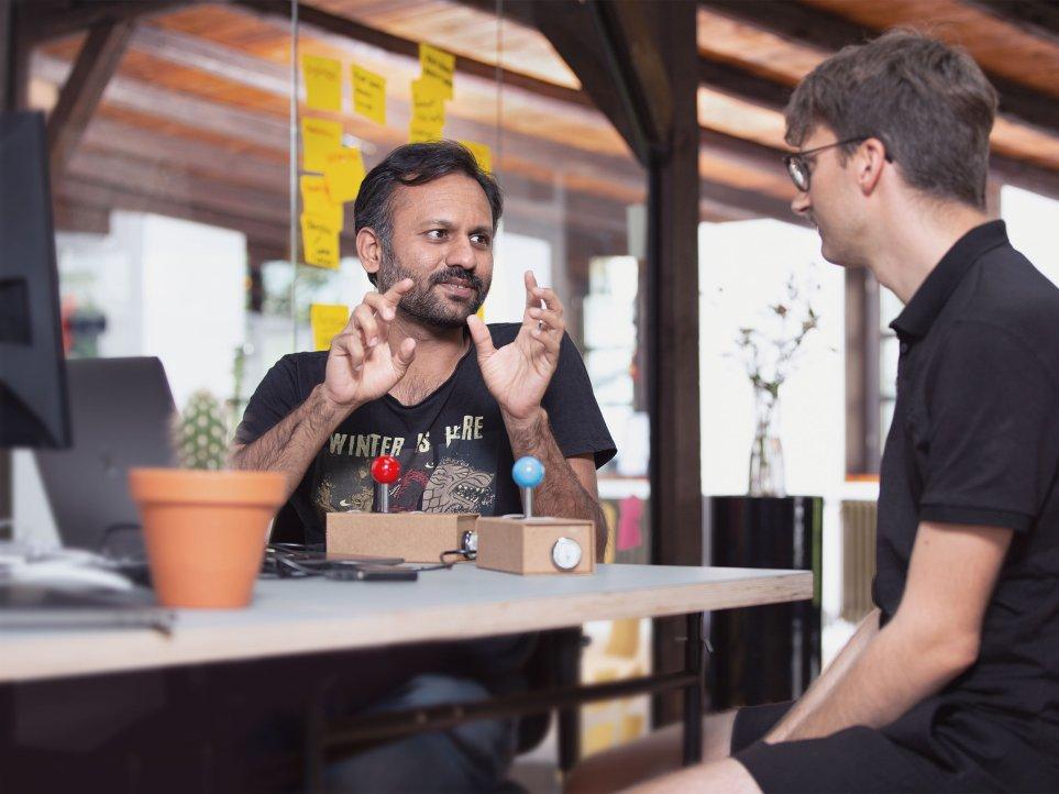 Shibin Das im Gespräch mit Julian an einem Tisch mit zwei Retro-Attari-Controllern.