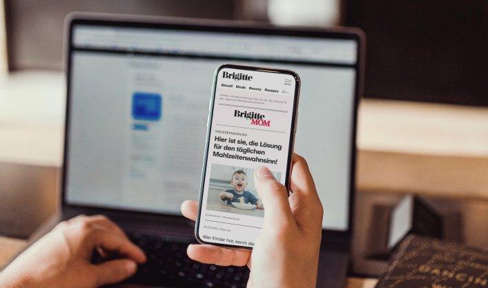 Eine Hand hält ein Smartphone, das die Brigitte Website zeigt. Im Hintergrund ist ein geöffneter Laptop zu sehen.