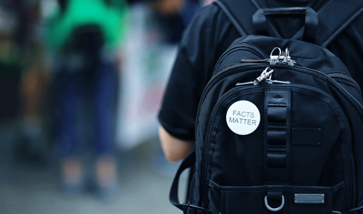 Person von hinten mit schwarzem Rucksack und weißem Abzeichen mit der Aufschrift "facts matter" in urbaner Umgebung.