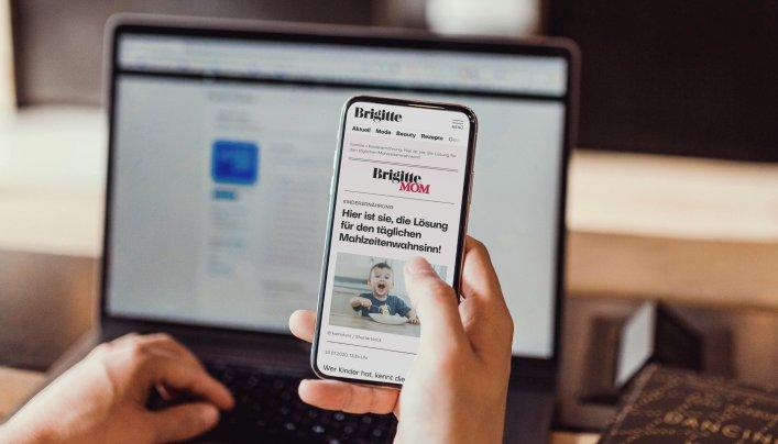 Eine Hand hält ein Smartphone, das die Brigitte Website zeigt. Im Hintergrund ist ein geöffneter Laptop zu sehen.