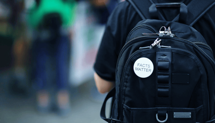 Person von hinten mit schwarzem Rucksack und weißem Abzeichen mit der Aufschrift "facts matter" in urbaner Umgebung.
