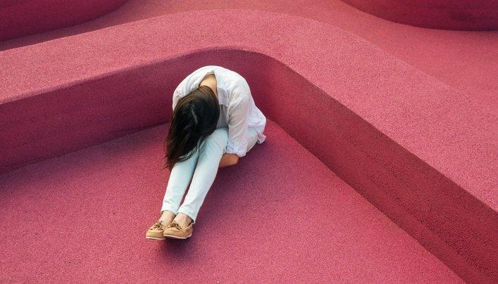 Frau sitzt allein mit gesenktem Kopf auf pinker Struktur – Sinnbild für seelische Krisen, mentale Belastung und den Bedarf an digitalen Hilfssystemen wie der TelefonSeelsorge.