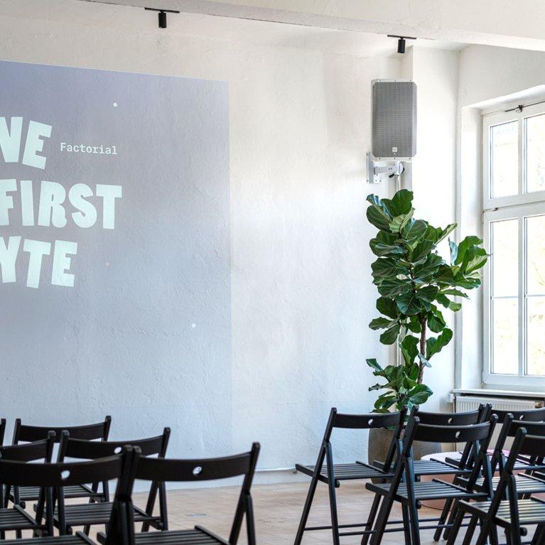 Moderner Eventraum bei Factorial mit Projektorwand „Love at First Byte“, hellen Fensterfronten, Pflanzen und bereitgestellten Stuhlreihen für Meetups oder Tech-Talks.