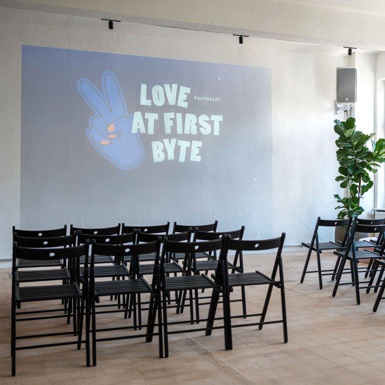 Moderner Eventraum bei Factorial mit Projektorwand „Love at First Byte“, hellen Fensterfronten, Pflanzen und bereitgestellten Stuhlreihen für Meetups oder Tech-Talks.