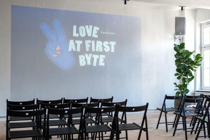Modern event space at Factorial featuring a “Love at First Byte” projector screen, bright window fronts, indoor plants, and neatly arranged chairs ready for meetups or tech talks.