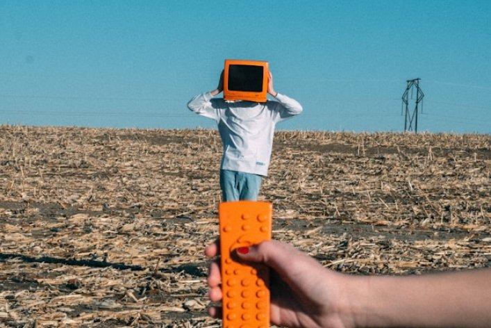 Eine Person in einem abgeernteten Feld mit orangefarbenem Monitor auf dem Kopf, während im Vordergrund eine Hand eine orange Fernbedienung auf sie richtet – ein Symbol für Kontrolle und Datensouveränität.