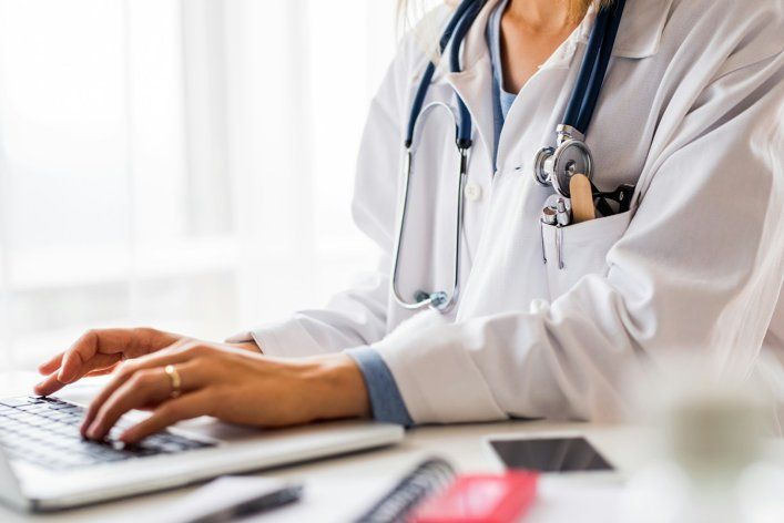 A woman in a white coat is sitting at a desk, working on her laptop.