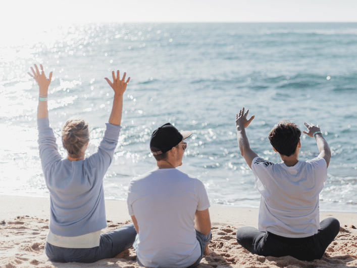 Image showing three of the Factorial colleagues sitting on the beach and facing the ocean. Their arms are raised to the sky