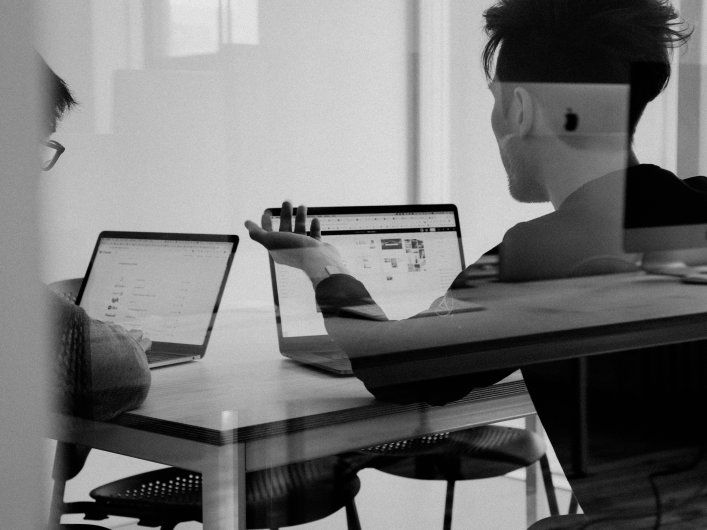 Kolleg*innen sitzen in einem Büro vor ihren Laptops und sprechen miteinander.